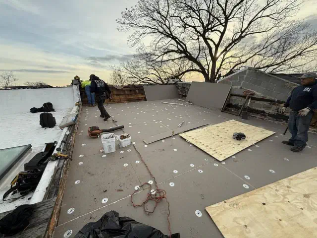Roofers fastening insulation boards and plywood onto a flat roof at sunset.
